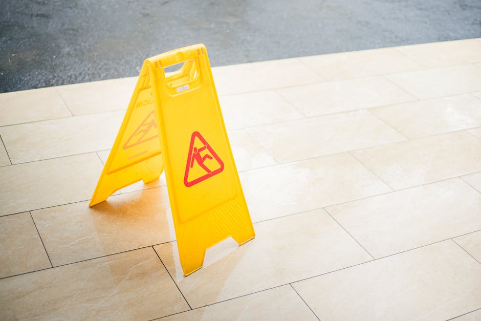 A yellow wet floor warning sign with a red triangle and an icon of a person slipping is placed on a light beige tiled surface outdoors. The tiles have a smooth, slightly glossy finish and are arranged