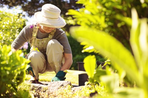 In the image, a woman wearing a wide-brimmed hat, beige trousers, and an apron is working outdoors in a well-maintained garden. She is kneeling on the grass and appears to be planting or tending to a flower bed, which is bordered by dark soil and edged with small stepping stones. Surrounding her are lush green plants and shrubs, with some taller foliage creating a natural backdrop. The garden features a neatly mown lawn area in the foreground, with dense, vibrant greenery and the sunlight casting warm highlights across the scene. The setting suggests a calm and tidy outdoor space, likely part of a residential garden in Putney, where gardening services from Gardener Putney could enhance landscape design or maintenance. The environment demonstrates a typical suburban garden layout with a mix of lawn, border plants, and shaded areas created by nearby trees, under clear weather conditions, contributing to an inviting and cared-for outdoor area.