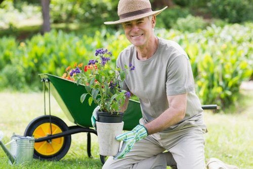 A smiling man wearing a wide-brimmed straw hat, light grey t-shirt, and garden gloves, kneeling on the lawn in a well-maintained outdoor space, holding a potted flowering plant with purple, red, and pink blooms. In the background, there is a lush green hedge, a vibrant grassy lawn, and garden trees providing shade. To the side, a bright yellow and black garden wheelbarrow is positioned on the grass, partially filled with gardening tools and supplies, indicating active outdoor maintenance. The scene is lit by natural daylight, suggesting a clear, mild weather day in a suburban garden setting, highlighting aspects of landscaping and gardening services available in Putney.